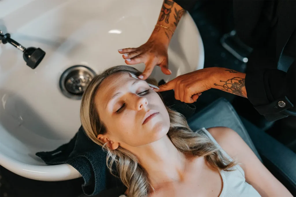 A person with closed eyes reclines at a salon sink while another person massages their temples with both hands, enjoying a moment of relaxation at an Aveda salon in Nashville. - True Blue Salon in Nashville, TN