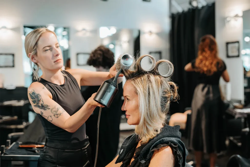 A hairstylist uses a blow dryer and large rollers to style a seated woman’s hair in a modern Aveda salon Nashville, specializing in haircuts and styling. - True Blue Salon in Nashville, TN