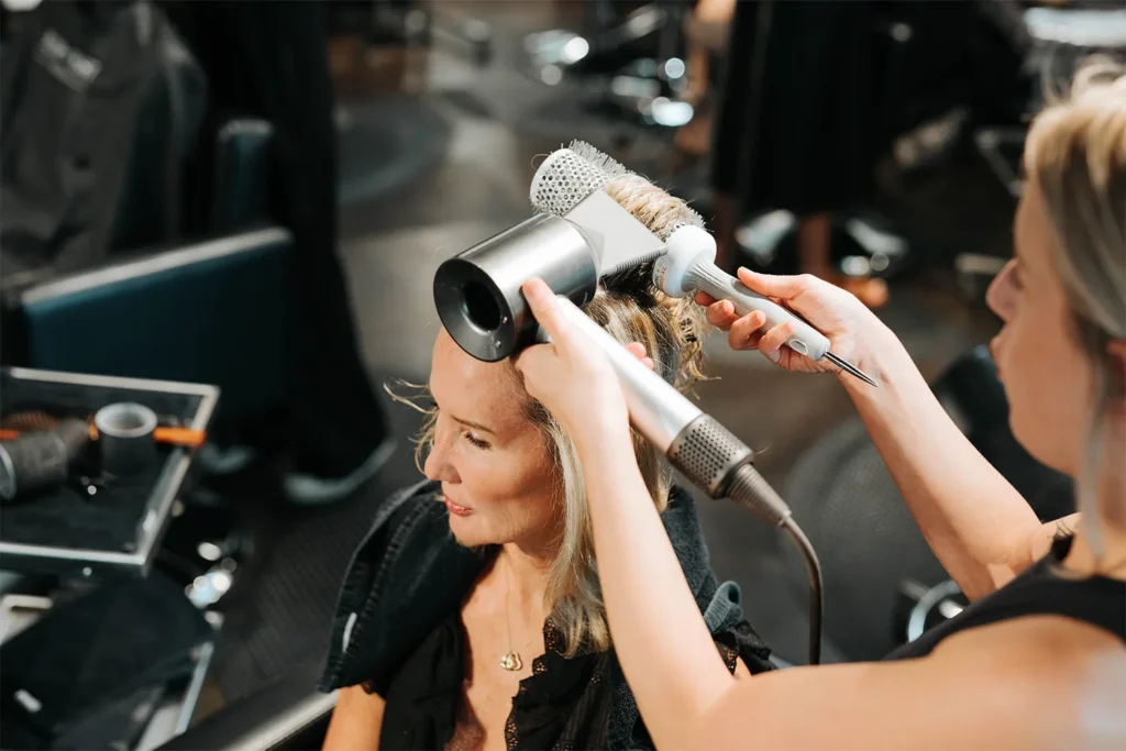 A woman is having her hair blow-dried and styled with a round brush by a hairstylist at an Aveda salon in Nashville, where clients enjoy expert services including hair color and highlights. - True Blue Salon in Nashville, TN