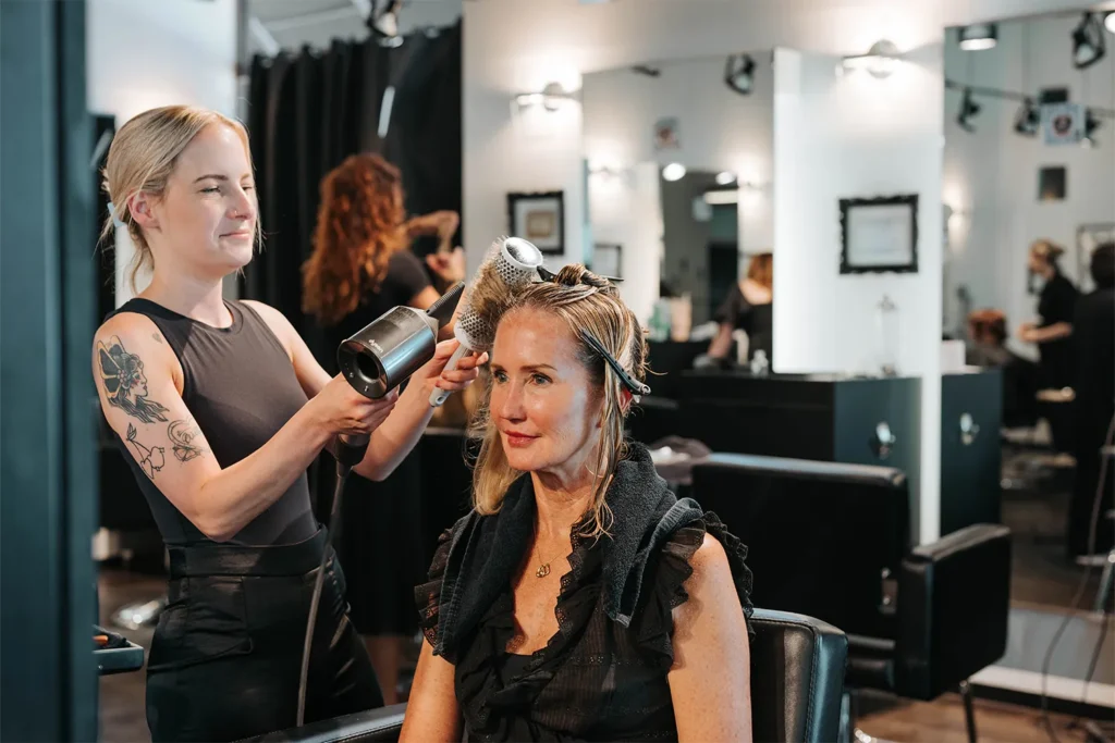 A hairstylist uses a round brush and blow dryer to style a seated woman’s hair in a modern salon, where clients can also enjoy expert hair color and highlights. Other clients and stylists are visible in the background. - True Blue Salon in Nashville, TN
