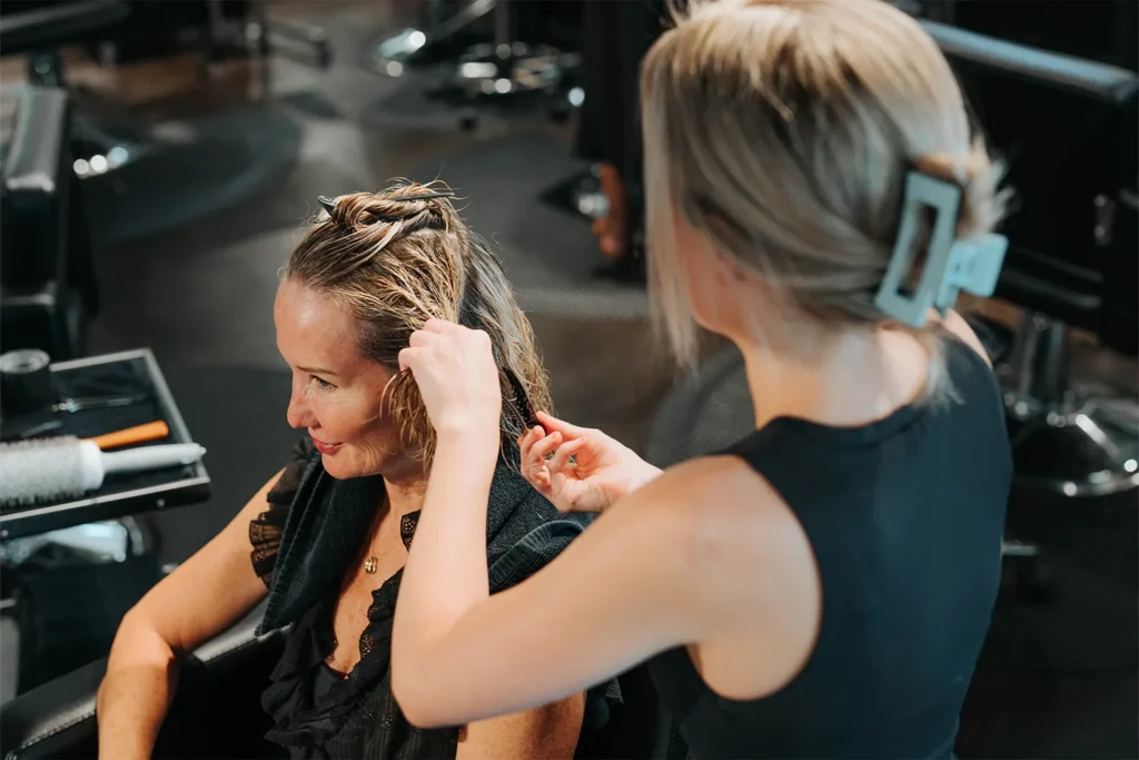 A hairstylist at an Aveda salon in Nashville is sectioning and combing a seated woman’s wet hair in a modern salon setting, where clients can also enjoy exclusive salon gift cards and specials. - True Blue Salon in Nashville, TN