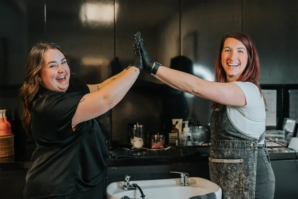 Two women in a Nashville hair salon give each other a high five and smile, standing next to a hair washing station with salon supplies visible in the background. - True Blue Salon in Nashville, TN