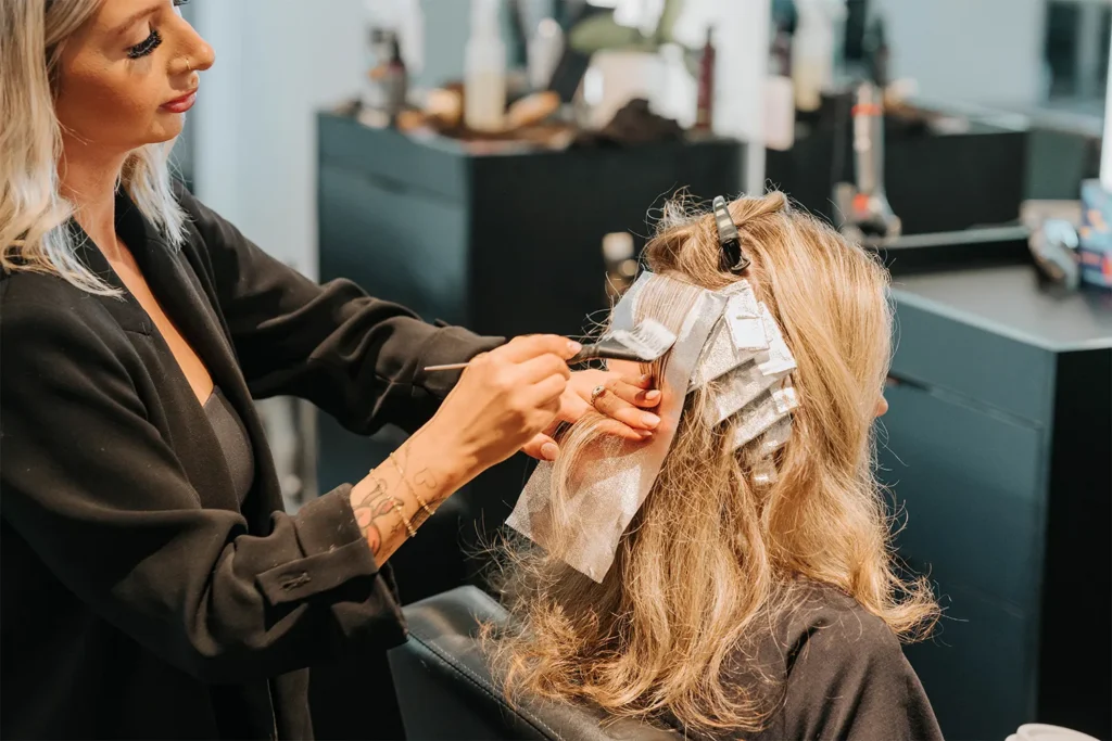 A hairdresser applies hair dye to a client’s hair using foil sections in a salon, expertly creating beautiful hair color and highlights. Various salon tools and products are visible in the background. - True Blue Salon in Nashville, TN