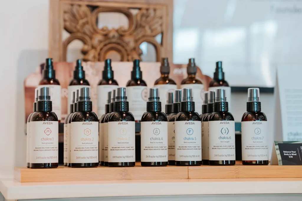 A row of Aveda branded spray bottles with different chakra numbers and colors displayed on a wooden tray, with more bottles in the background, captures the tranquil ambiance of our Nashville hair salon. Discover premium hair color and highlights services while enjoying this thoughtfully curated space. - True Blue Salon in Nashville, TN