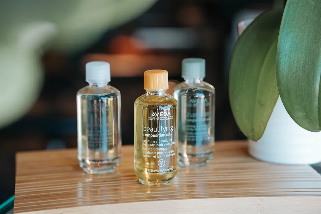 Three small bottles of oil, including Aveda Beautifying Composition Oil, are arranged on a wooden surface next to a green plant—an inviting scene reminiscent of the curated ambiance found in a Nashville hair salon. - True Blue Salon in Nashville, TN