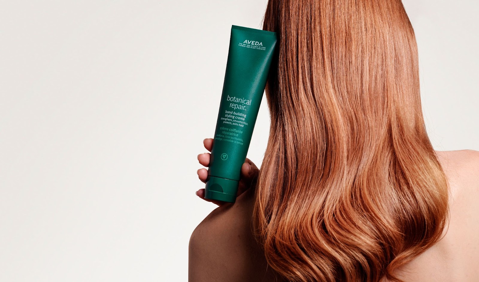 A person with long, wavy, beautiful red hair holds a green tube of Aveda Botanical Repair Intensive Strengthening Masque beside their head, showcasing the product. - True Blue Salon | Nashville, TN