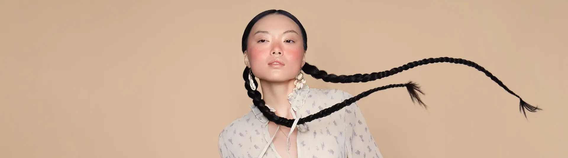 A woman with long, braided hair wears a patterned blouse and large earrings, posing in front of a beige background. - True Blue Salon | Nashville, TN