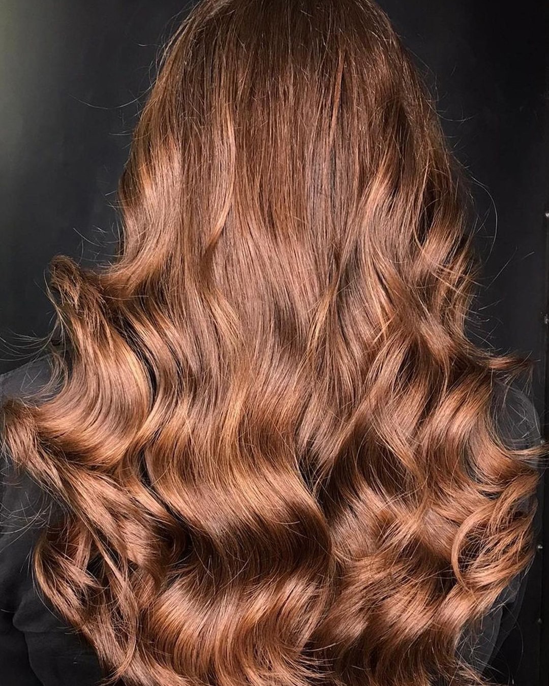 Back view of wavy, auburn hair against a dark background. - True Blue Salon | Nashville, TN