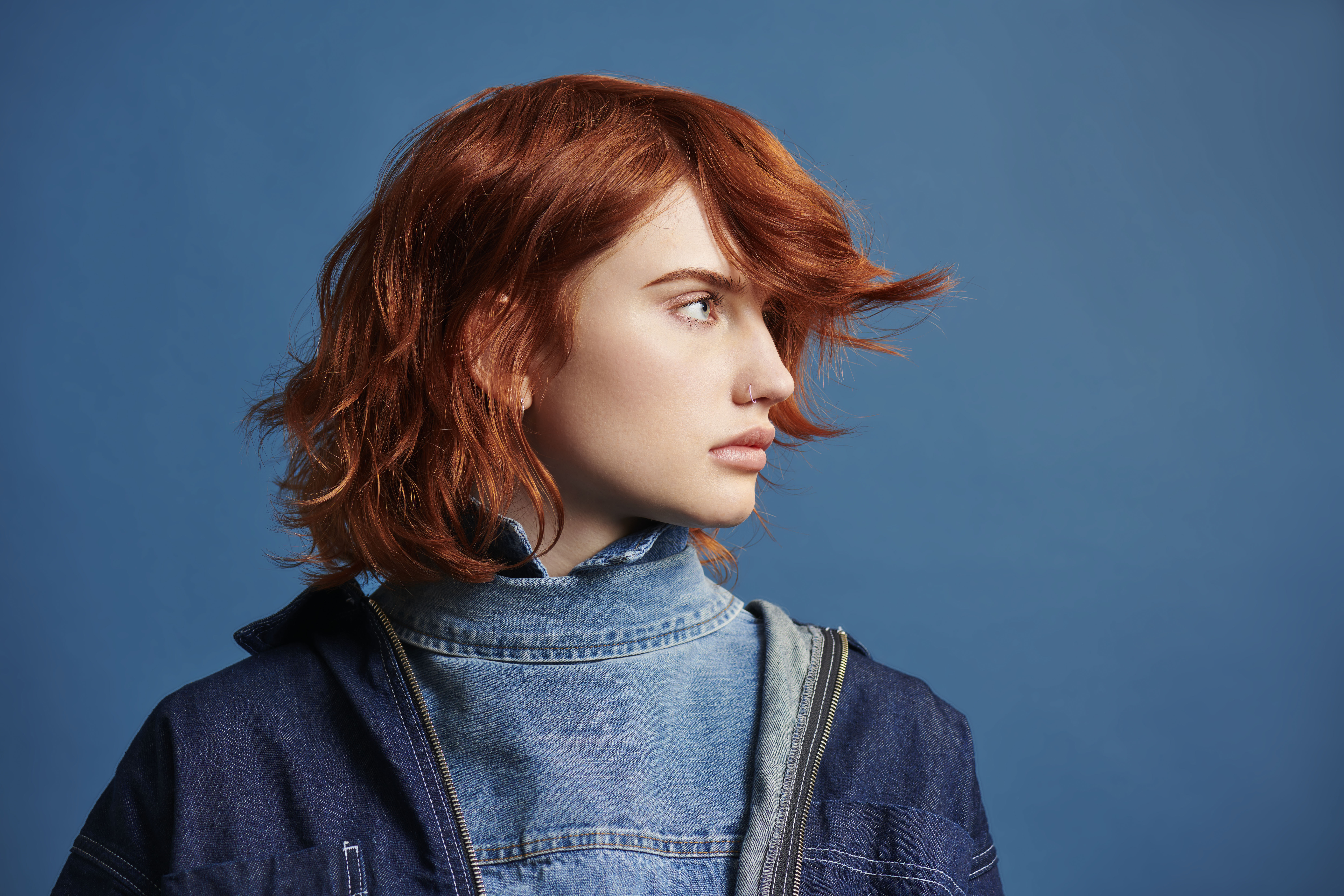 A person with red hair looks to the side against a blue background, wearing a layered denim and dark jacket. - True Blue Salon | Nashville, TN