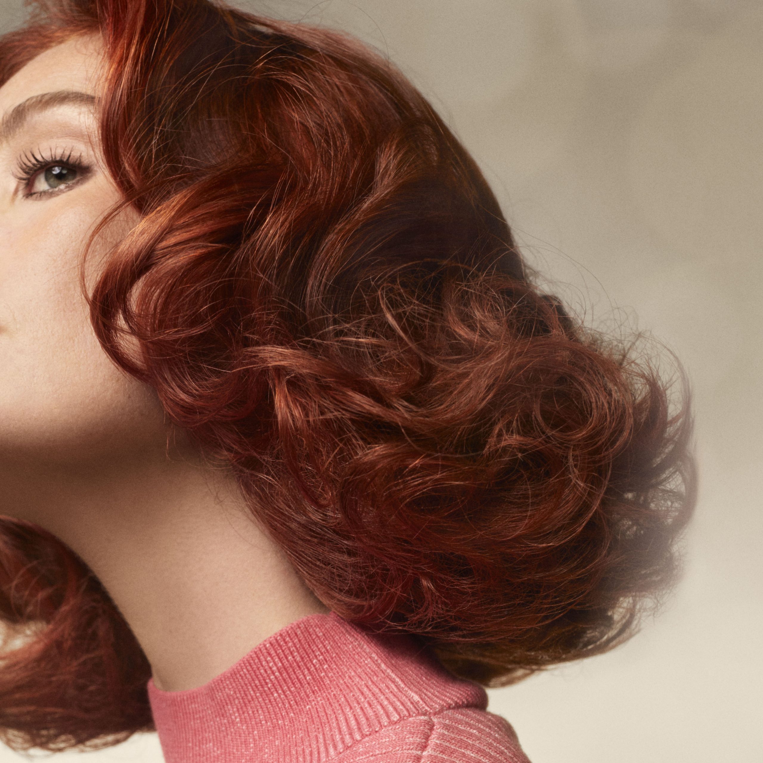 A person with wavy red hair, wearing a pink top, is pictured from the side against a soft, neutral background. - True Blue Salon | Nashville, TN