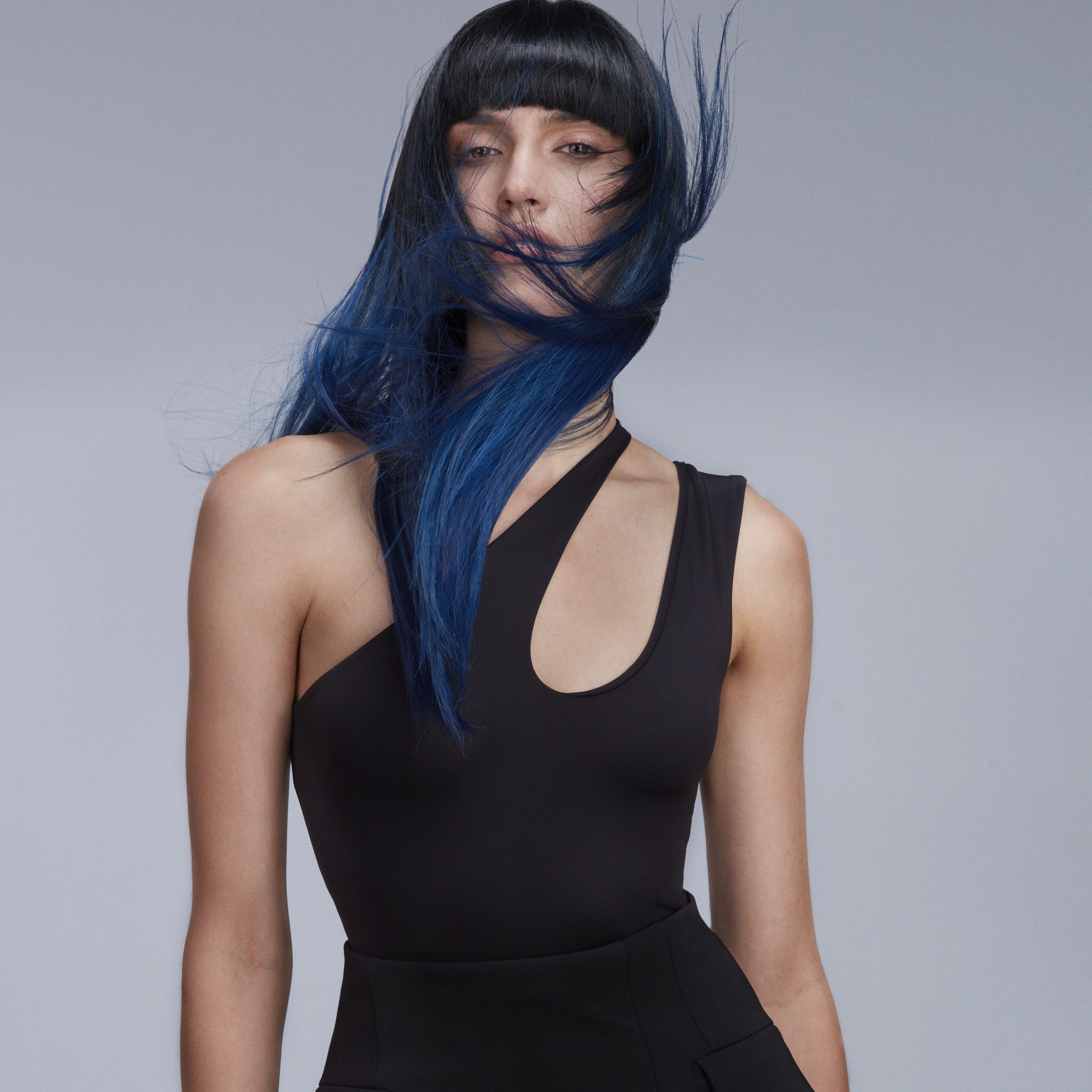 Person with long, blue-tinted hair and bangs wears a black asymmetrical top against a plain background. Hair partially obscures face. - True Blue Salon | Nashville, TN