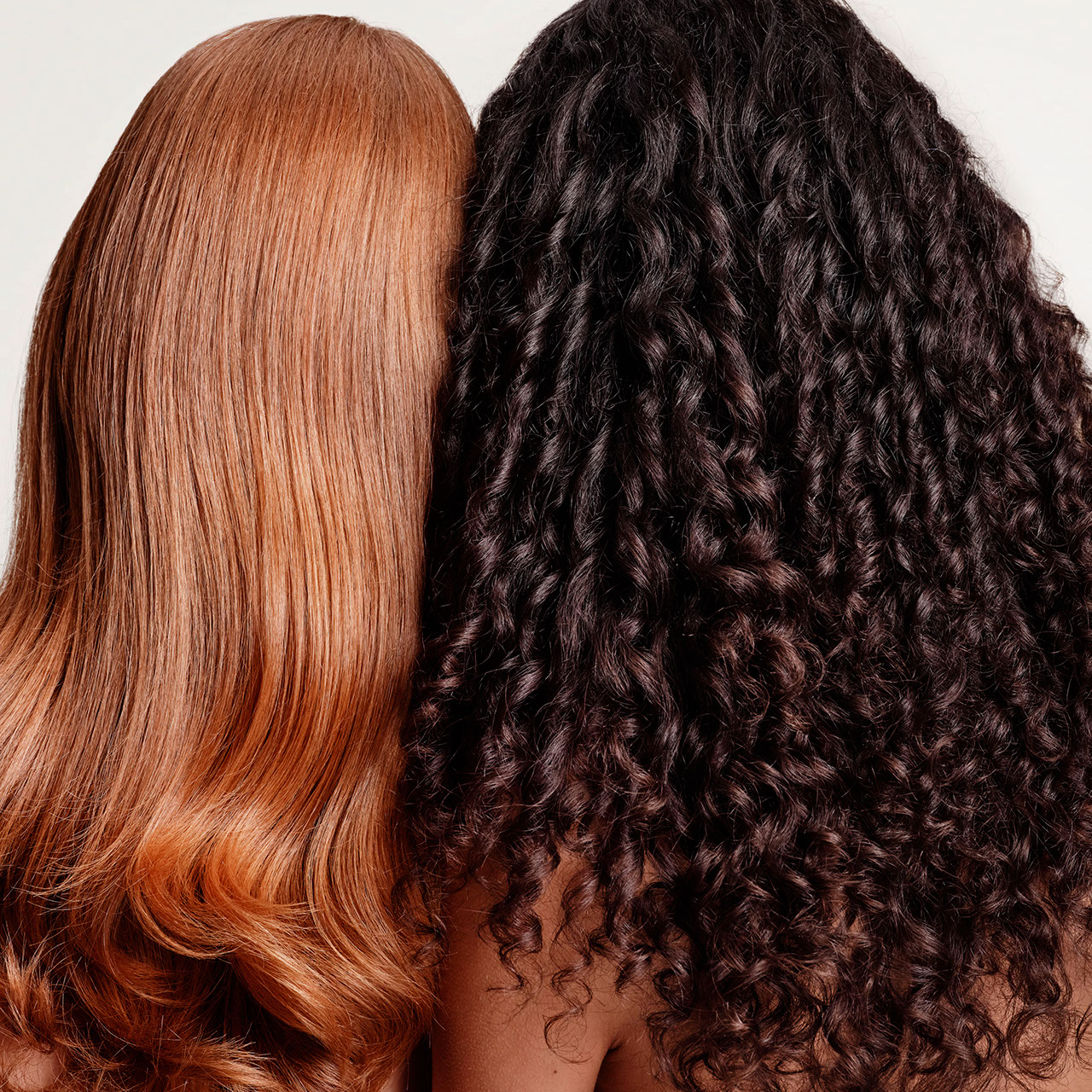 Two people with long hair from the back: one with straight red hair, the other with curly dark brown hair. Their heads are close together against a plain background. - True Blue Salon | Nashville, TN