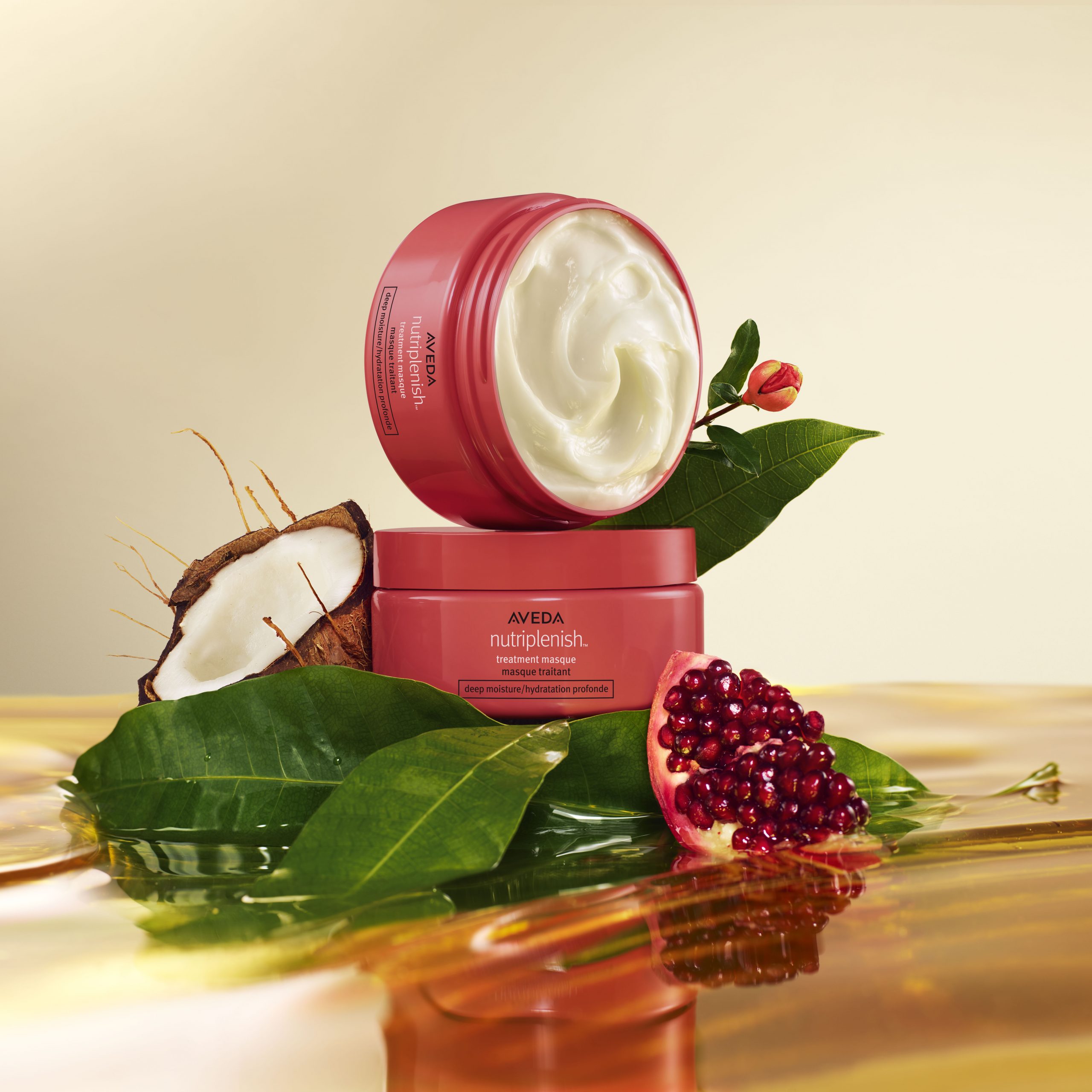 Aveda Nutriplenish hair mask containers with pomegranate seeds and coconut on leaves, set against a glossy golden surface. - True Blue Salon | Nashville, TN