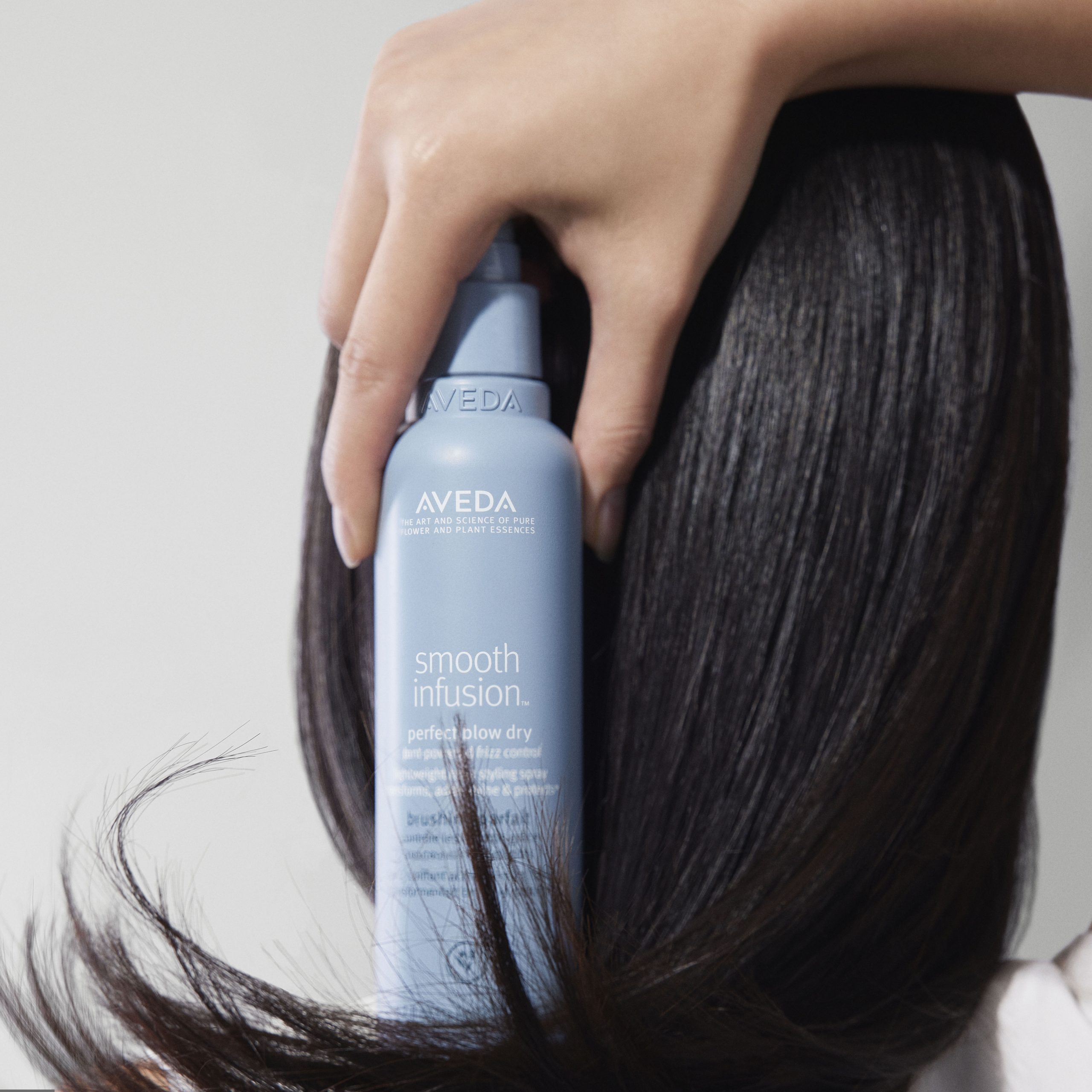 A hand holds a blue bottle of Aveda Smooth Infusion hair product against straight, black hair. - True Blue Salon | Nashville, TN