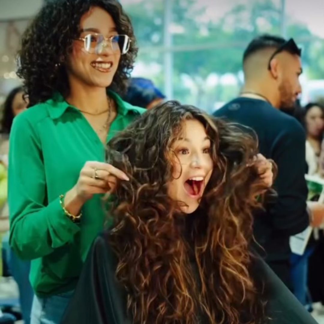 A woman with curly hair sitting in a salon chair looks excited as a stylist stands behind her, holding her hair and smiling. - True Blue Salon | Nashville, TN