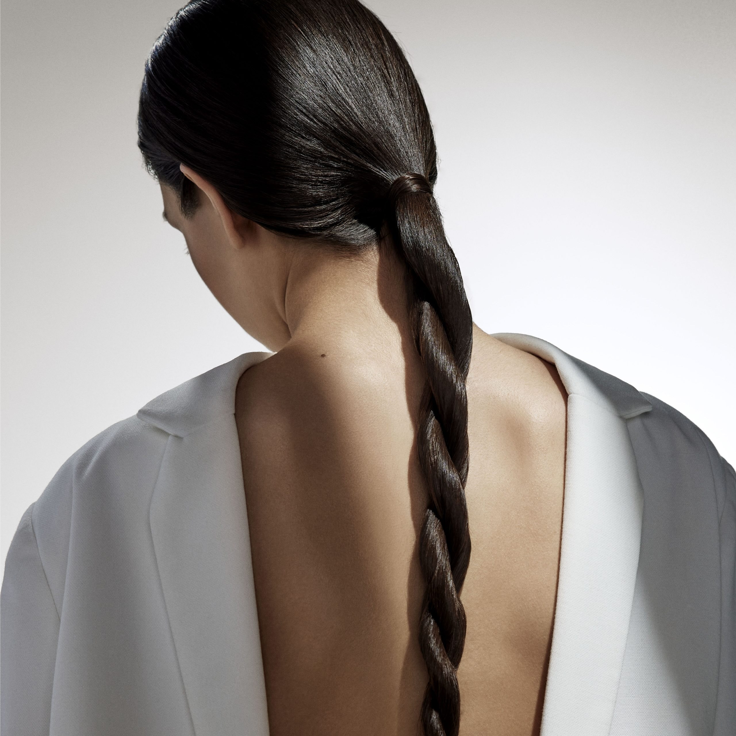 A person with long dark hair styled in a braid, wearing a white garment with an open back, is shown from behind against a neutral background. - True Blue Salon | Nashville, TN