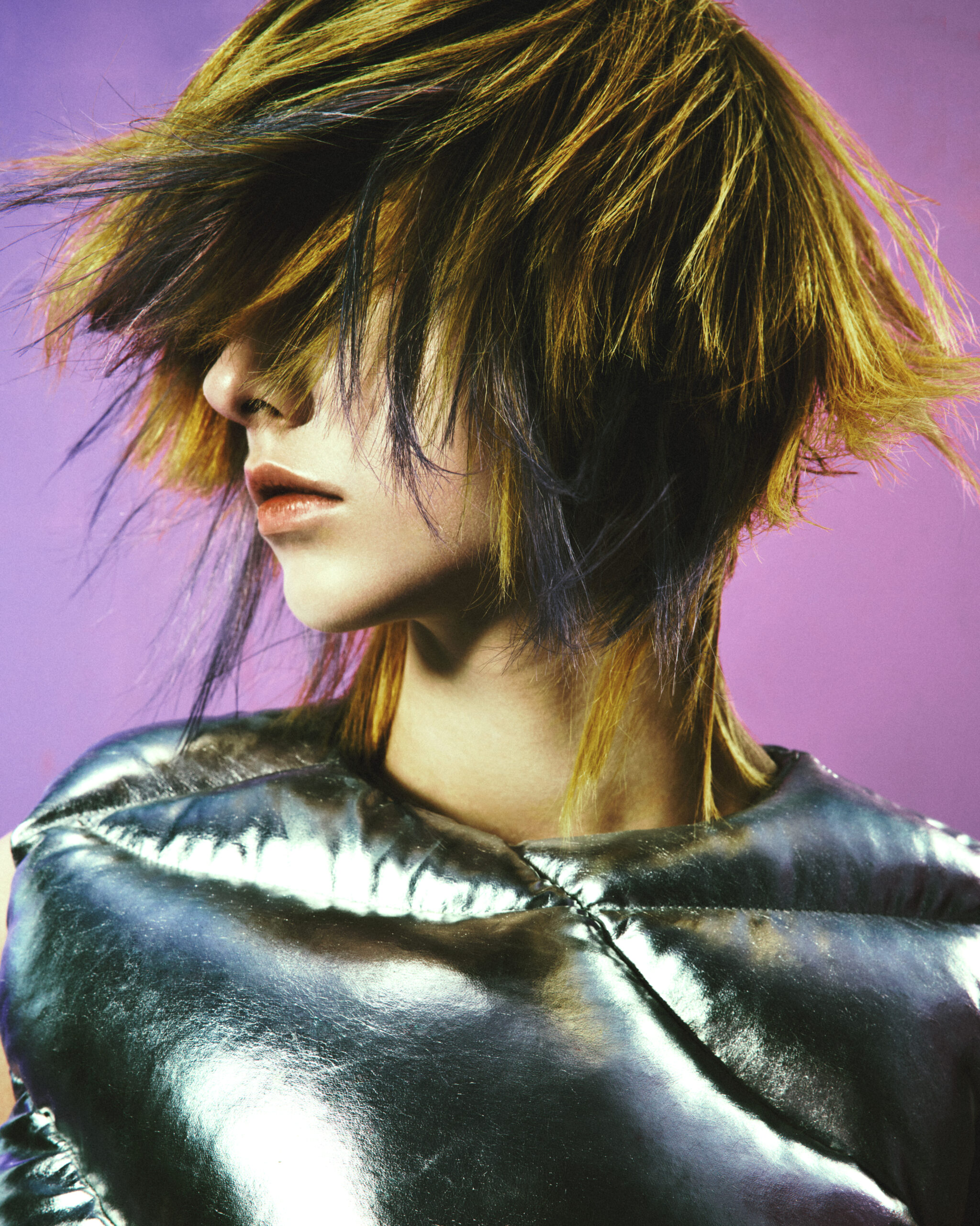 Person with shaggy, highlighted hair that beautifully showcases expert hair color and highlights, dressed in a metallic silver top, poses against a purple background, looking to the side with hair partially covering their face. - True Blue Salon in Nashville, TN